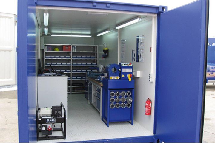 Blauer Container, der als Werkstatt eingerichtet ist. Im Inneren befinden sich Regale mit Werkzeugen und Ersatzteilen, eine Werkbank mit Maschinen, eine Feuerlöscherstation und ein kleiner Stromgenerator auf dem Boden. An der Rückwand sind weitere Werkzeuge und Zubehör ordentlich gelagert.  